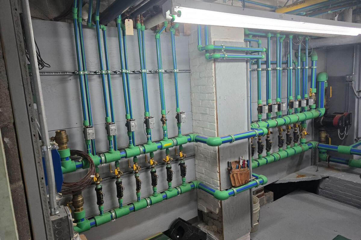 A mechanical room featuring an organized array of green and blue plastic piping with valves, gauges, and control modules mounted on the wall. The system is cleanly installed and labeled, running vertically and horizontally around a painted brick column, with tools hanging neatly in a pouch.