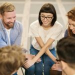A group of six people sit in a circle, smiling and talking, with some holding hands or touching arms in a supportive gesture.