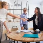 Two women shake hands across a round table in a modern office, while a man stands nearby. Laptops, documents, and coffee cups are on the table.