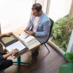 Two people sit at a small table in an office, one looking at documents and the other using a smartphone, with notebooks and a tablet on the table.